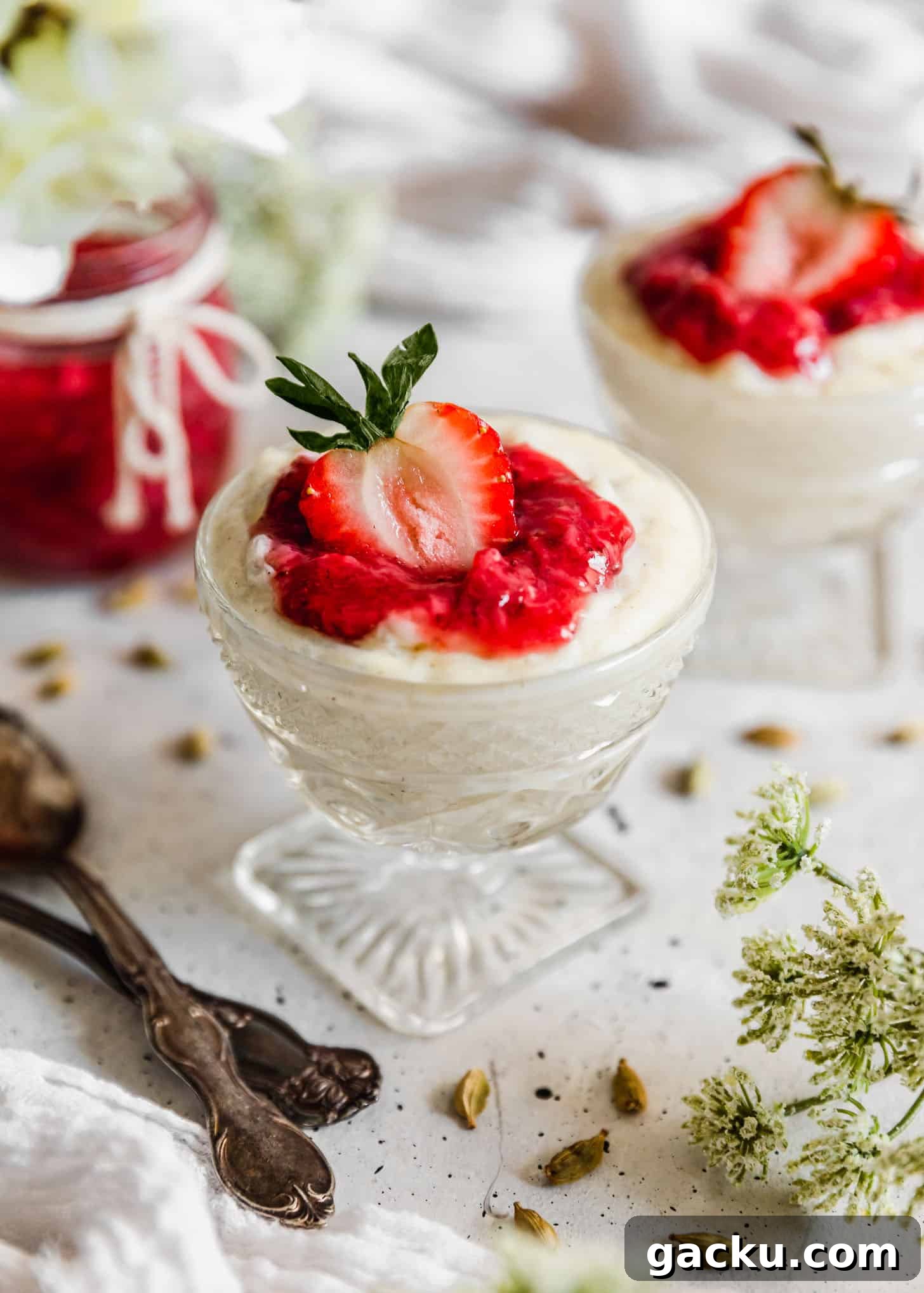 Creamy Cardamom Rice Pudding Recipe — Silky Indian Kheer Guide 6 Two glass dishes of cardamom rice pudding with strawberries next to cardamom pods, a jar of jam, spoons, and white blossoms on a white counter.