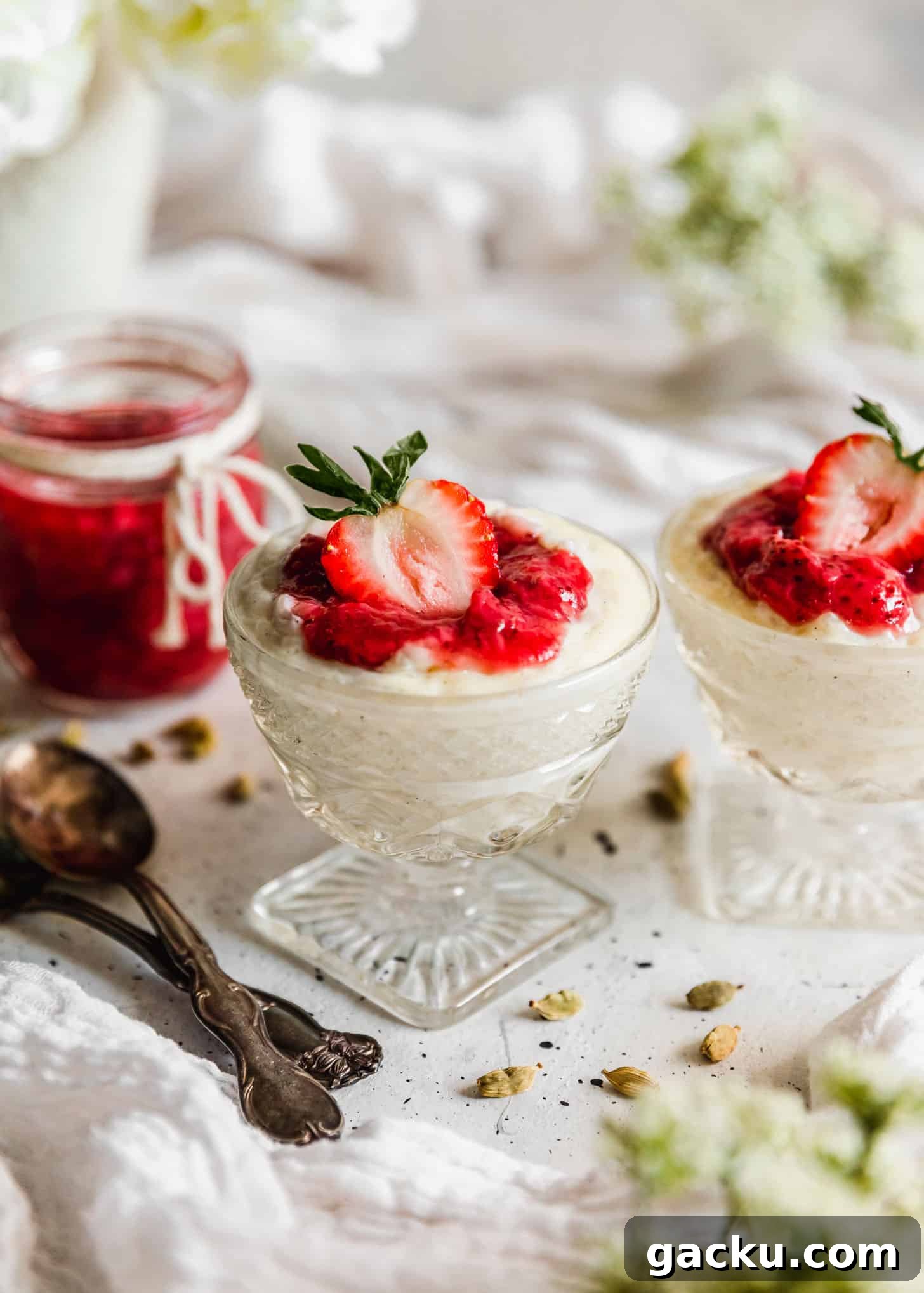 Creamy Cardamom Rice Pudding Recipe — Silky Indian Kheer Guide 7 Two glass dishes of cardamom rice pudding with strawberries next to jam, spoons, cardamom pods, and white flowers on a white table.