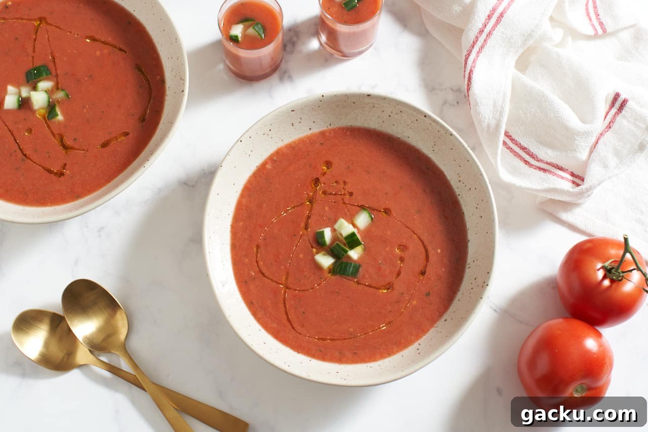Two bowls of gazpacho and two shot glasses of gazpacho topped with chopped cucumber. Two gold spoons, two tomatoes, and a red and white striped towel surround the bowls.