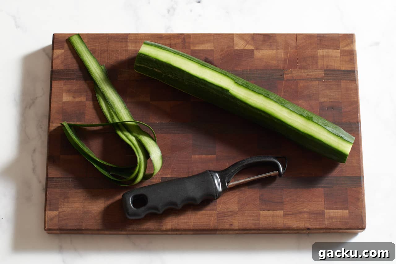 A vegetable peeler on a wooden cutting board with a cucumber that has strips of peel removed.