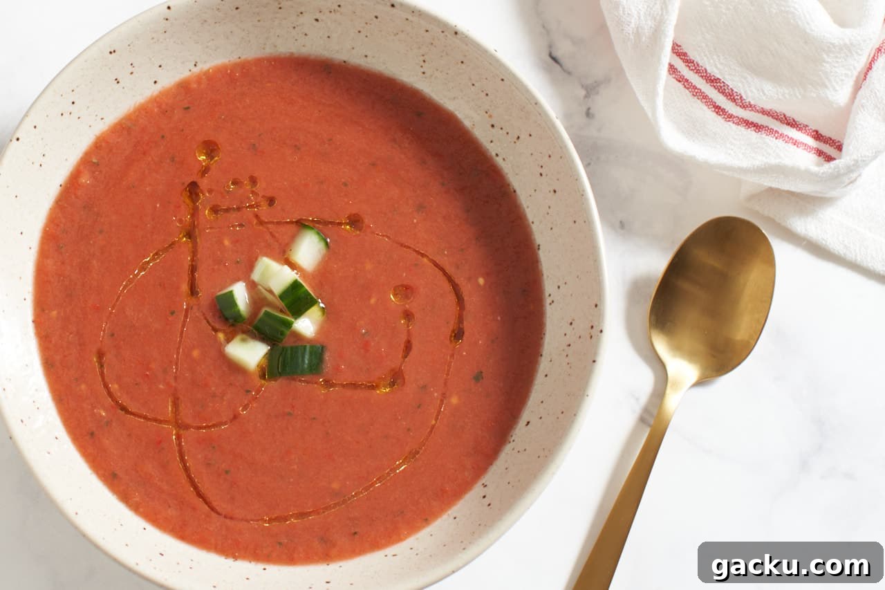 A bowl of gazpacho topped with chopped cucumber and olive oil next to a gold spoon and a red and white striped towel.