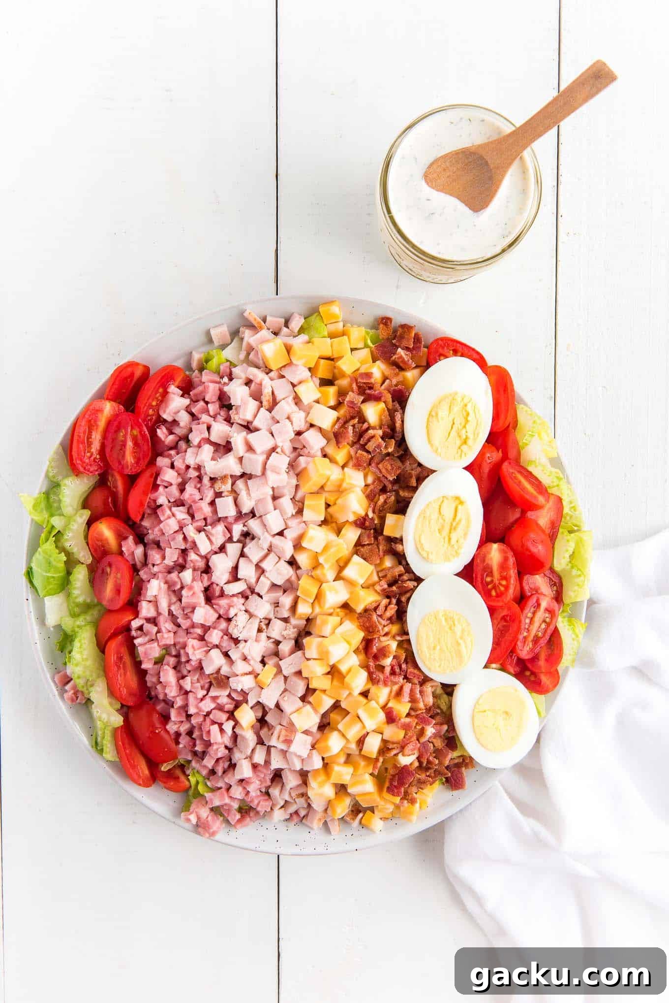 A Chef salad with rolls of turkey, ham, cheese, bacon, eggs, tomatoes, and celery with a jar of ranch dressing.