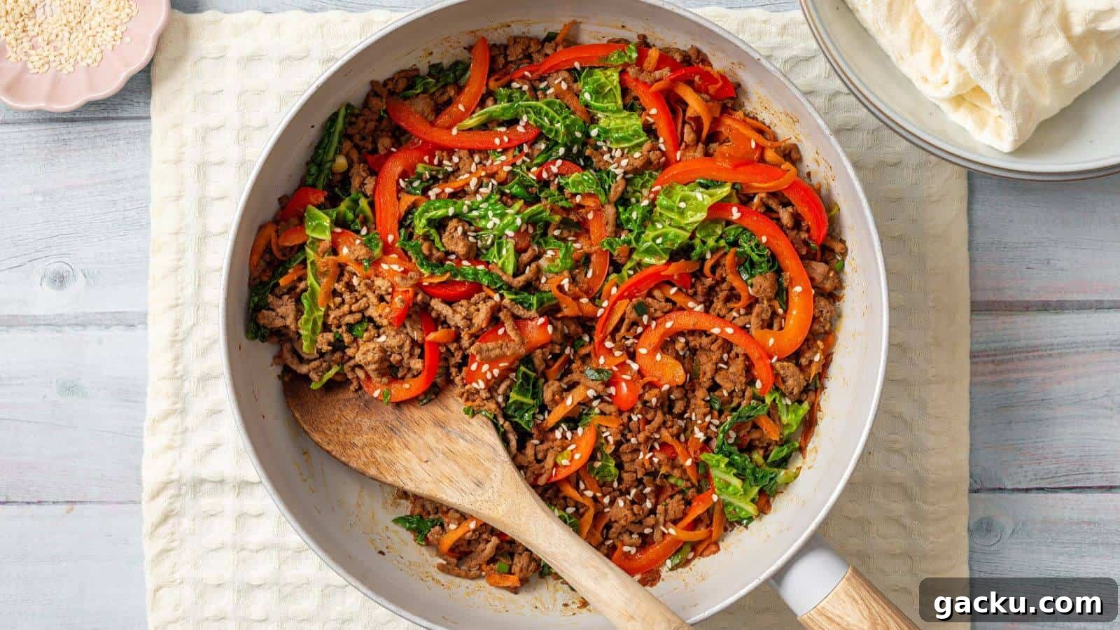 30 One-Pan Skillet Meals for Quick Cleanup and Big Flavor 27 A skillet containing a stir-fried mix of ground meat, sliced bell peppers, leafy greens, and white sesame seeds, with a wooden spoon resting on the side. Plates with flatbreads are in the background.
