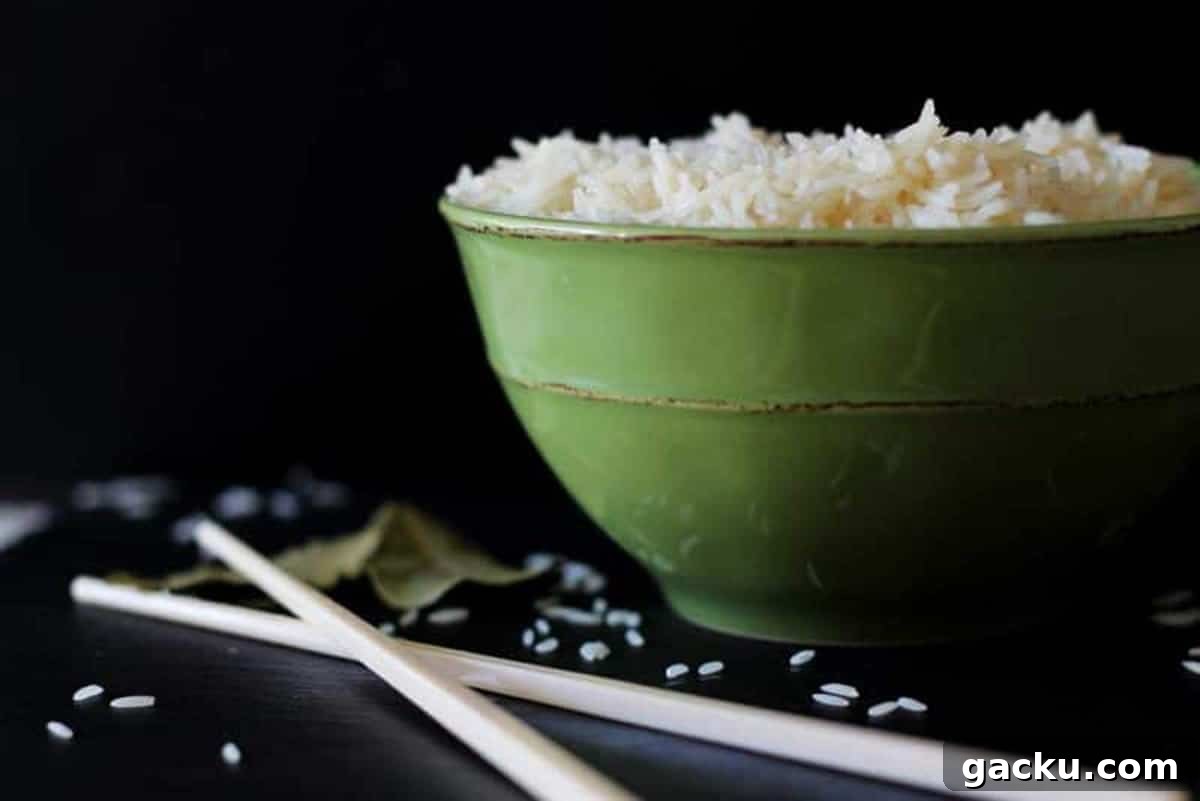 Simple Rice Pilaf Recipe: Fluffy, Flavorful Rice Pilaf in 30 Minutes 9 Green bowl sitting on a black table containing white rice, crossed chopsticks, bay leaf and rice granules.