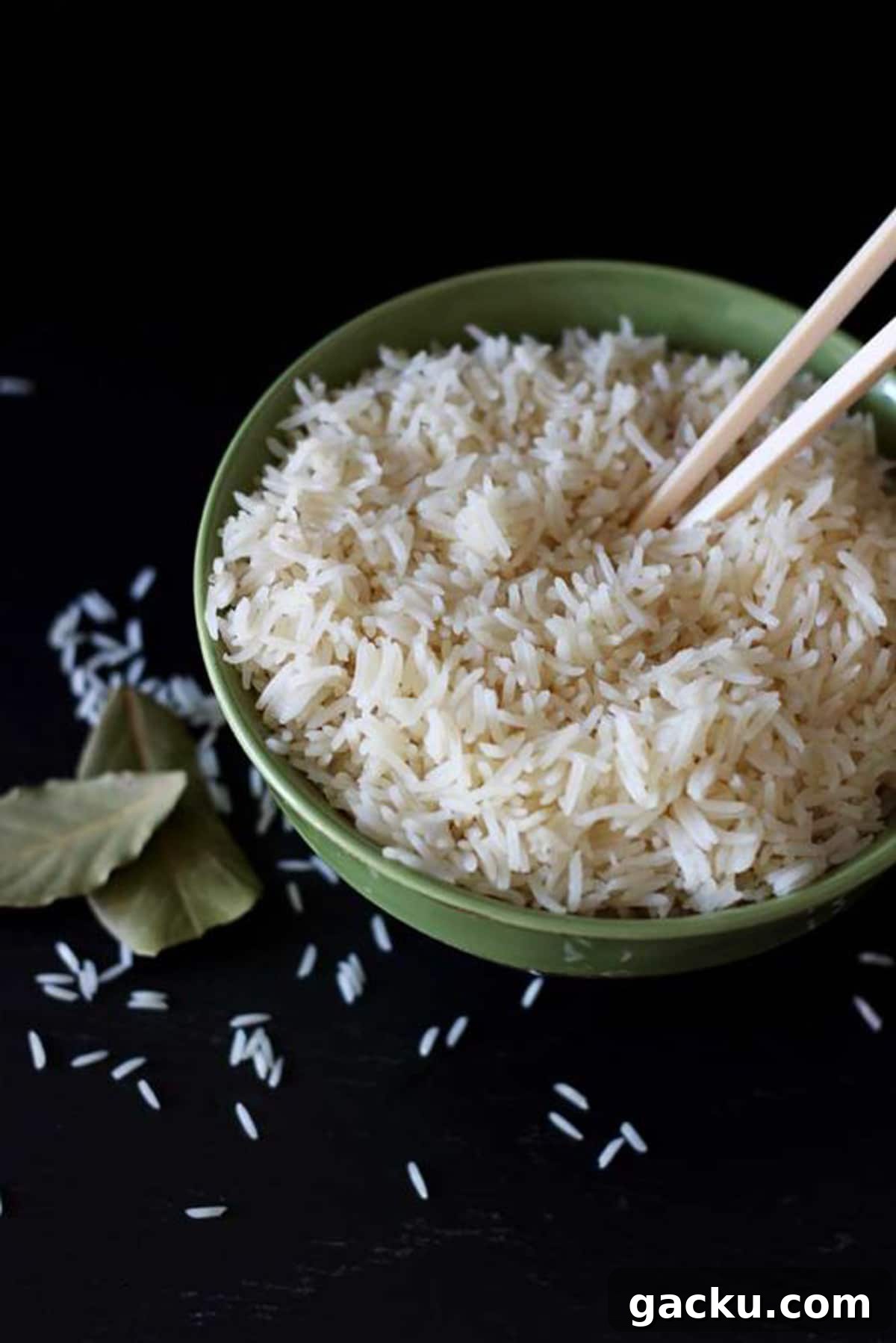 Simple Rice Pilaf Recipe: Fluffy, Flavorful Rice Pilaf in 30 Minutes 3 Green bowl sitting on a black table containing white rice with chopsticks in bowl, rice granules and bay leaf on table.