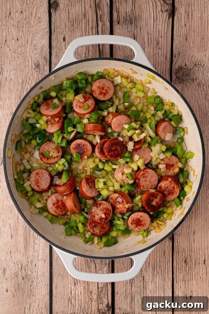 Diced onion, green bell pepper, and celery sautéing in a skillet with browned sausage rounds.