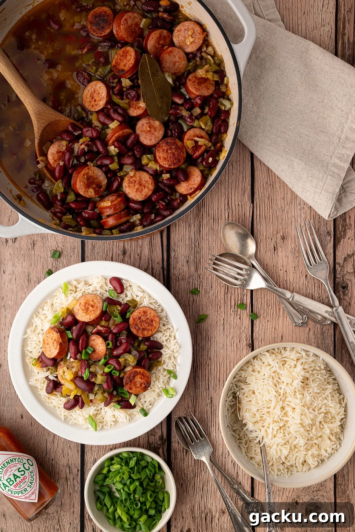 A white bowl filled with fluffy white rice topped with a generous ladle of red beans and smoked sausage, garnished with sliced green onions and Tabasco sauce.