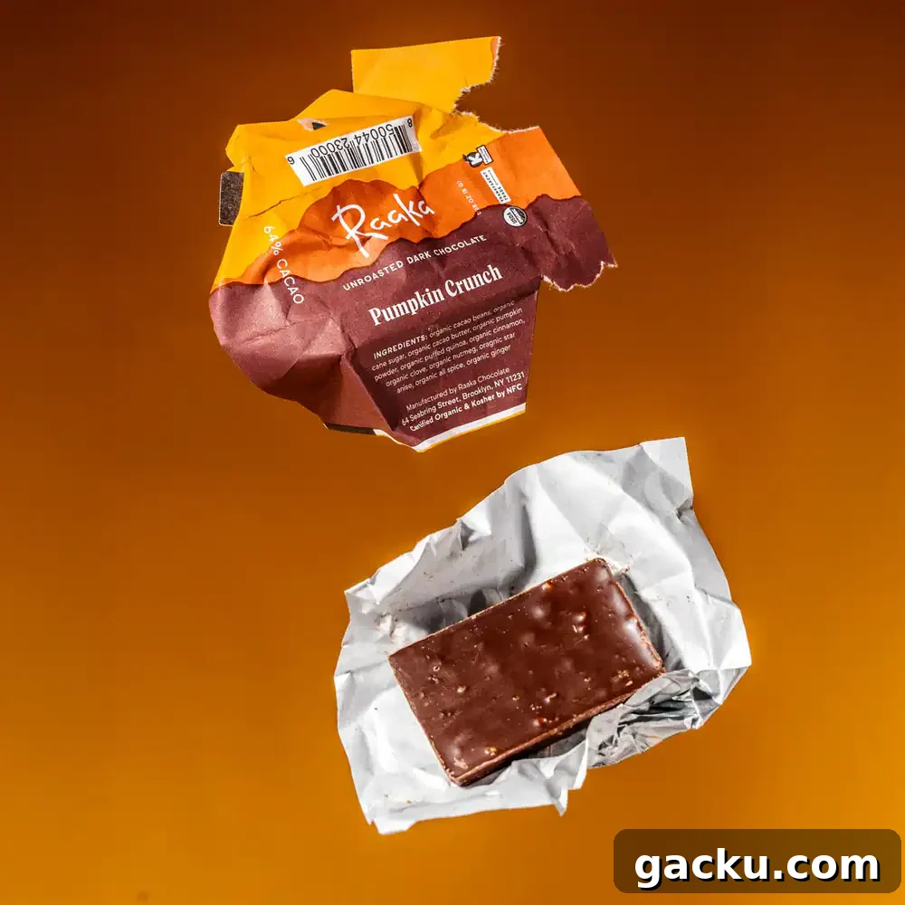 A half-eaten Raaka Pumpkin Crunch chocolate bar is shown on a crumpled silver wrapper, with a torn orange wrapper above it, all on a solid orange background.