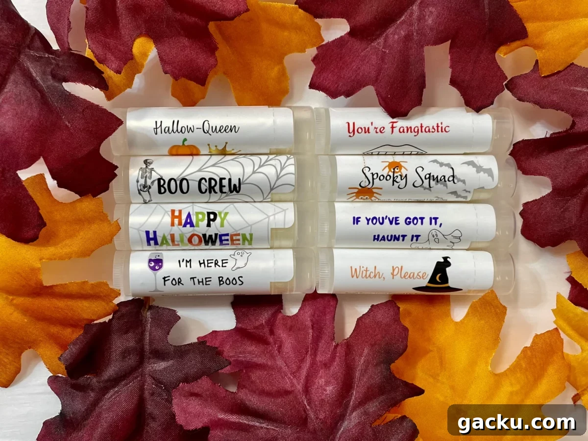 Eight lip balm tubes with Halloween-themed labels are arranged in two columns on a white surface and bordered by artificial autumn leaves in shades of red, burgundy, and orange.