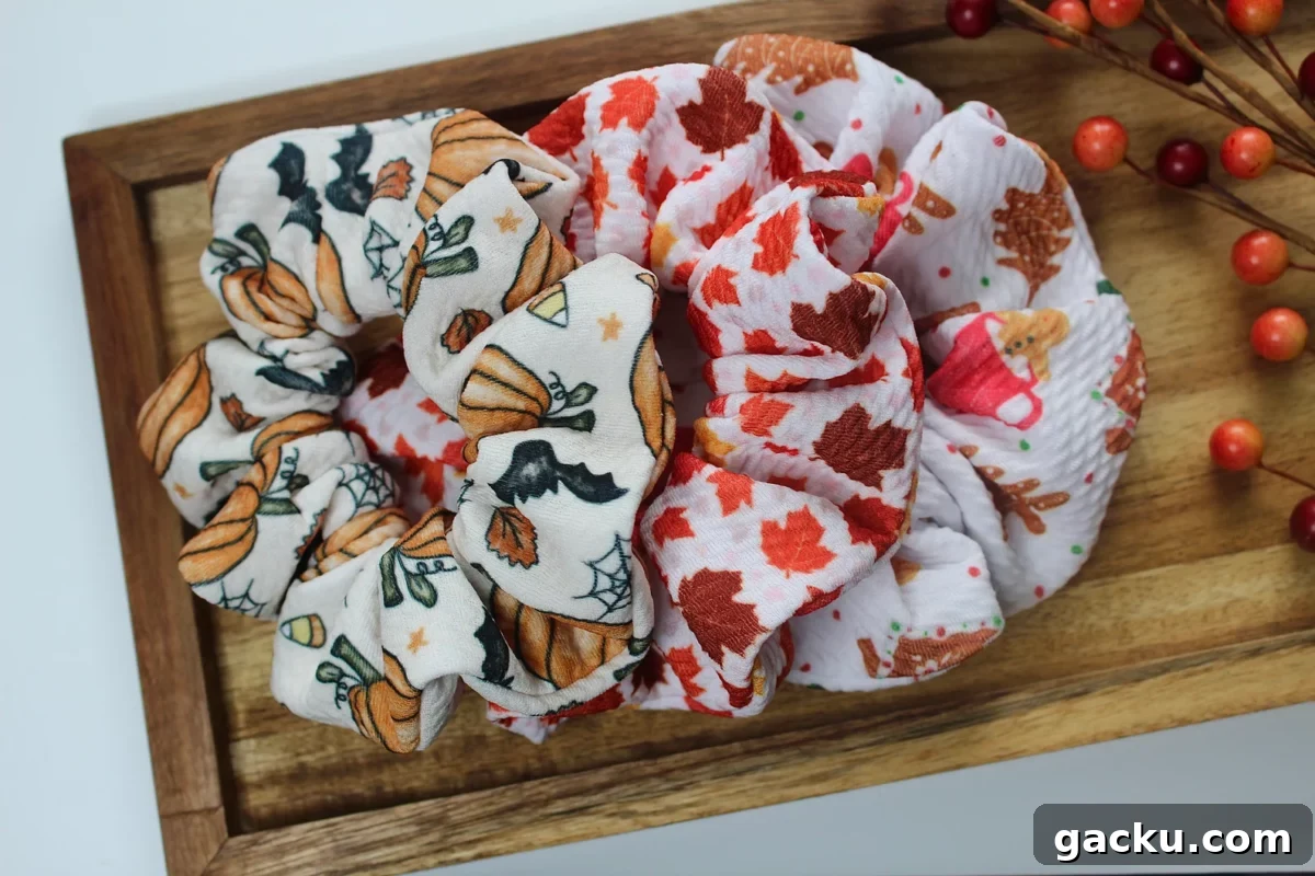 An overhead shot displays two fall-themed scrunchies—one with pumpkins and bats, and one with maple leaves—lying in a rustic wooden tray with berry sprigs in the top right corner.