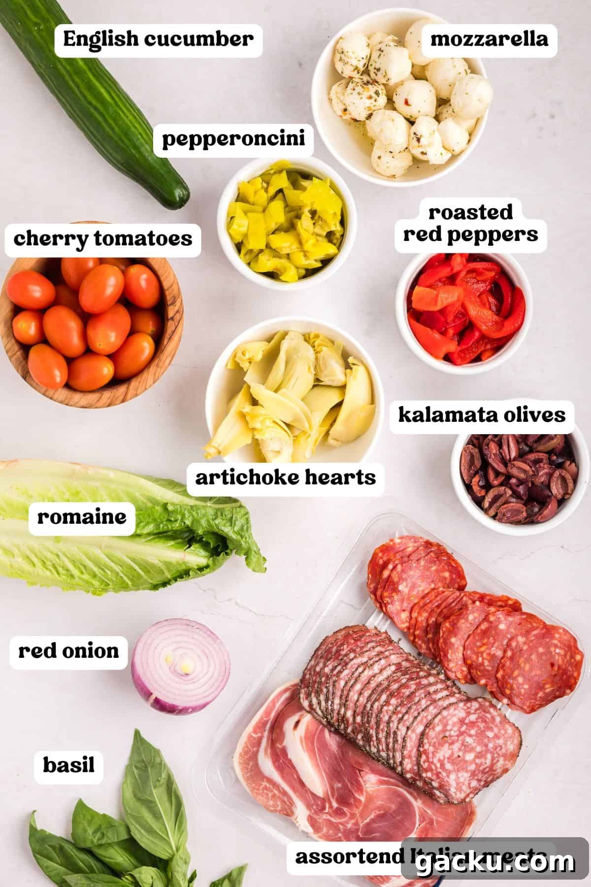 Flat lay of antipasto salad ingredients, including labeled mozzarella, pepperoncini, cherry tomatoes, roasted red peppers, kalamata olives, artichoke hearts, cucumber, red onion, romaine, basil, and assorted Italian meats.