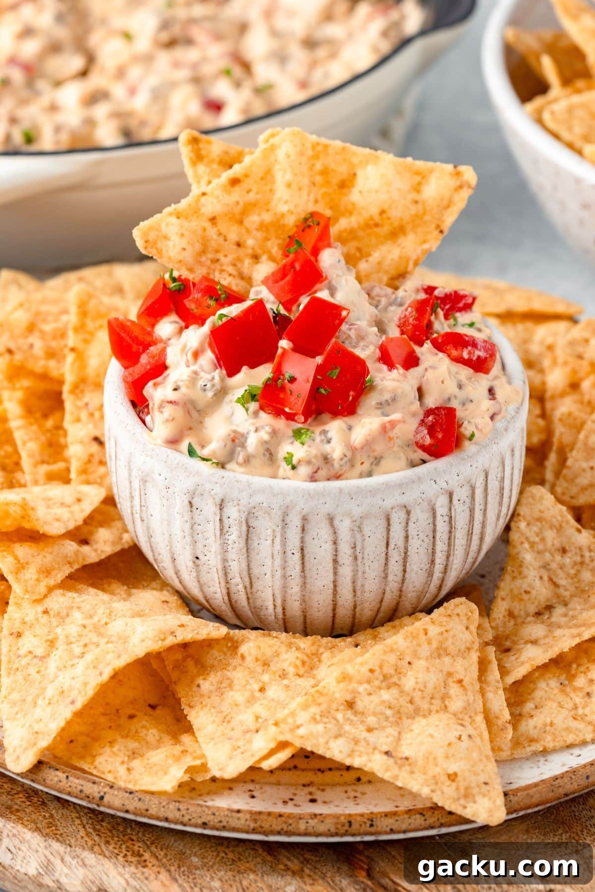 A small white bowl filled with creamy dip topped with diced tomatoes and herbs is surrounded by a pile of tortilla chips on a plate. The background shows more chips and a larger bowl of dip.