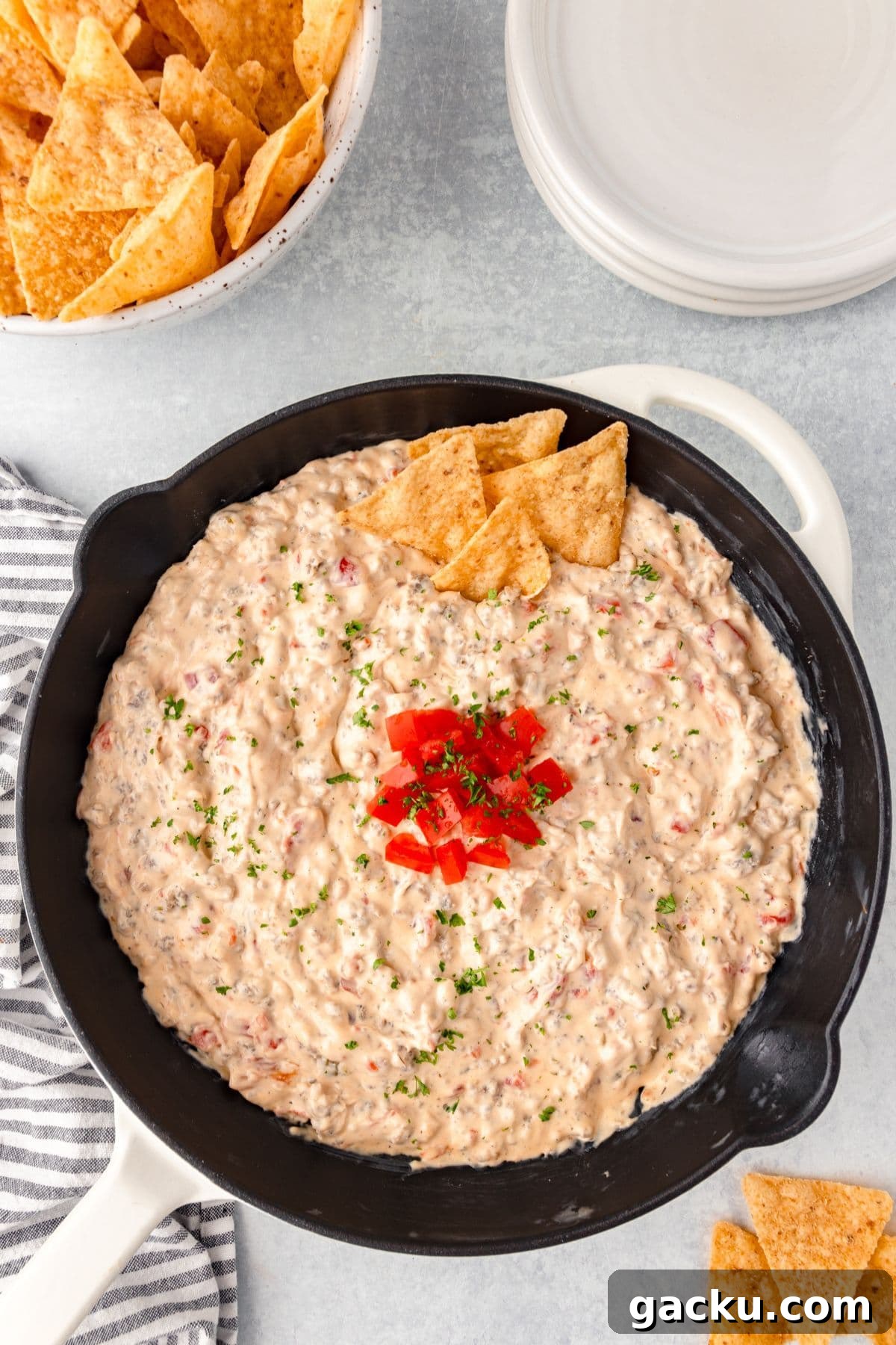 A cast iron skillet filled with creamy dip, garnished with chopped tomatoes and herbs, and accompanied by triangular tortilla chips. A bowl of chips and a stack of plates are in the background on a light surface.